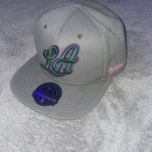 Grå keps från Mitchell & Ness - Snygg grå keps med broderad 'LA' logga i grönt och lila framtill. Märket Mitchell & Ness är broderat i rosa på sidan. Kepsen har en platt skärm och är i storlek 7 1/8. Pris kan diskuteras vid köp!