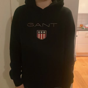 Svart hoodie från GANT - Säljer en stilren svart hoodie från GANT med deras ikoniska logga och ett sköldemblem på bröstet. Perfekt för en avslappnad och bekväm stil. Hoodien har långa ärmar och en klassisk passform. Hör av er vid funderingar!