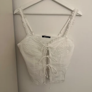 Vit knytblus från Gina Tricot - Säljer en söt vit blus från Gina Tricot med knytning framtill och spetsdetaljer. Blusen har smala axelband och en fin struktur i tyget. Perfekt för en somrig look!