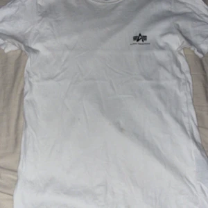 Vit t-shirt från Alpha Industries - Säljer en klassisk vit t-shirt från Alpha Industries. Den har en enkel design med korta ärmar och en liten logga på bröstet. Perfekt för en avslappnad stil.