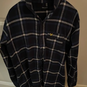 Rutig skjorta från Lyle & Scott - Snygg rutig skjorta från Lyle & Scott i mörkblått och vitt. Skjortan har långa ärmar och en klassisk krage. Den är dekorerad med ett gult broderat emblem på bröstfickan. Perfekt för en stilren look.