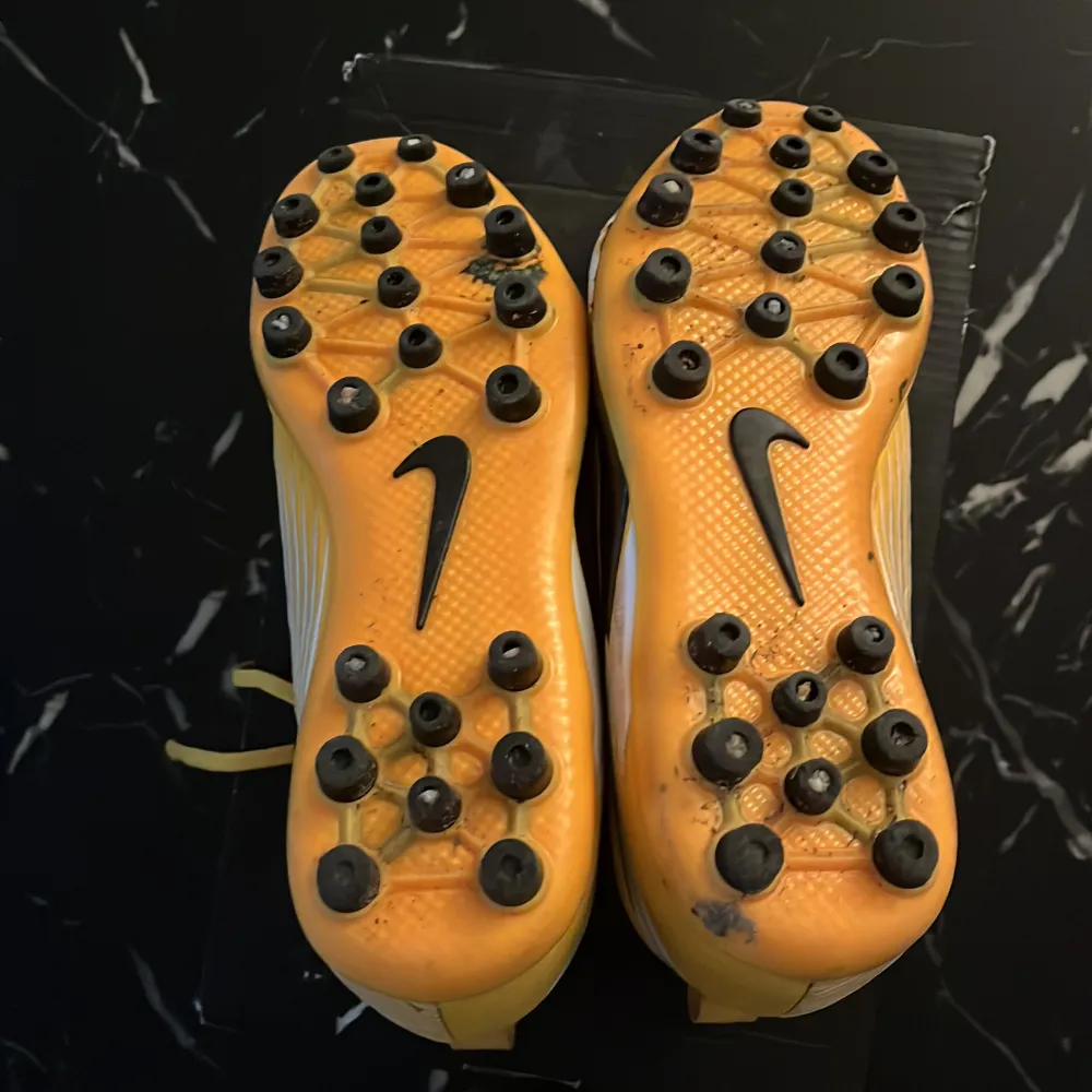 Snygga gula fotbollsskor från Nike med svart och vit detalj på sidan. Skorna har snörning och är designade för optimal hastighet på planen. Perfekta för fotbollsspelare som vill sticka ut. Den är även signerad av Omar Faraj, Bojan Djordjic och Jardell Kanga.. Kengät.