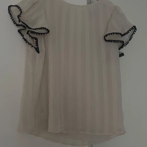 Vit blus med svarta volangdetaljer - Charmig vit blus med korta ärmar och volangdetaljer. Blusen har ett subtilt randigt mönster och är dekorerad med svarta kanter på ärmarna. Perfekt för en stilren och elegant look.