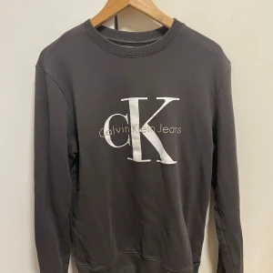 Calvin Klein Jeans tröja - Stilren och bekväm långärmad tröja från Calvin Klein Jeans i mörk färg med stor CK-logga på bröstet. Perfekt för en casual men trendig look. Storlek S 
