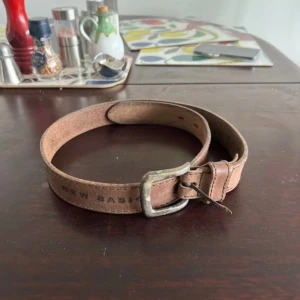 Brunt bälte från Sand - Säljer ett stilrent brunt bälte från Sam med texten 'SAND NEW BASIC' tryckt längs remmen. Bältet har ett klassiskt metallspänne och flera hål för justering. Perfekt för att komplettera din outfit.