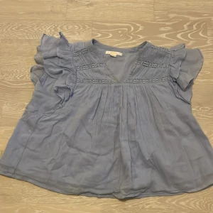 Blå blus från Gina Tricot - Säljer en söt blå blus från Gina Tricot med volangdetaljer på ärmarna. Blusen har en luftig passform och v-ringad hals. Perfekt för varma dagar!