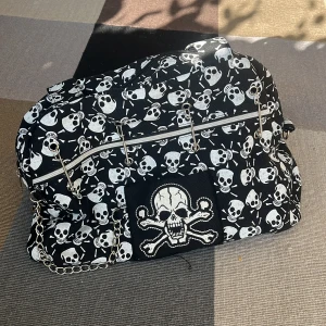 Väska - Säljer denna väska med dödskallemotiv! 🖤 Svart med vita dödskallar, säkerhetsnålsdetaljer och kedjor. Rymlig och praktisk för vardagsbruk eller som statement-accessoar. Är i oanvänt skick!