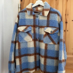 Rutig overshirt från SensUniq - Säljer en superfin rutig overshirt från SensUniq i blått, brunt och beige. Den har en avslappnad passform med långa ärmar och stora fickor framtill. Perfekt för höst och vår när du vill ha något stilrent och bekvämt. Knapparna framtill ger en klassisk look.