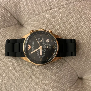 Emporio Armani klocka med svart armband - Säljer en snygg Emporio Armani klocka med ett svart armband och guld detaljer. Klockan har används ett fåtal gånger. Äkta i fint skick.
