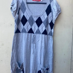 Grå stickad klänning med diamantmönster - Säljer en superfin grå stickad klänning med ett klassiskt diamantmönster i mörkblått och ljusgrått upptill. Klänningen har korta ärmar och en söt knytning i midjan för en justerbar passform. Perfekt för höst och vinter, och den har även praktiska fickor med dekorativa knappar. Passar både till vardags och festligare tillfällen!