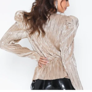 Glittrig beige blus från NLY Trend - Säljer en superfin glittrig beige blus från NLY Trend. Den har en v-ringad design och långa ärmar med puffiga axlar. Perfekt för festliga tillfällen! Materialet är glansigt och ger en lyxig känsla.