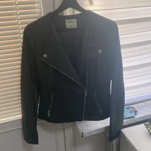 Svart skinnjacka från DeFacto - Säljer en snygg svart skinnjacka från DeFacto i storlek M. Jackan har en cool asymmetrisk dragkedja och stilrena knappar. Perfekt för höst och vår, och den ger en edgy look till vilken outfit som helst. Jackan är långärmad och i bra skick. Passar perfekt för både vardag och fest!