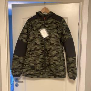 Säljer en snygg och praktisk camouflagejacka från Helly Hansen. Jackan har en cool camo-design med svarta detaljer på ärmarna och axlarna. Den är långärmad och har en dragkedja framtill. Perfekt för höst och vår, och den är i nyskick. Passar bra för utomhusaktiviteter och har en normal passform.