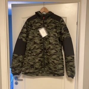Camouflagejacka från Helly Hansen - Säljer en snygg och praktisk camouflagejacka från Helly Hansen. Jackan har en cool camo-design med svarta detaljer på ärmarna och axlarna. Den är långärmad och har en dragkedja framtill. Perfekt för höst och vår, och den är i nyskick. Passar bra för utomhusaktiviteter och har en normal passform.