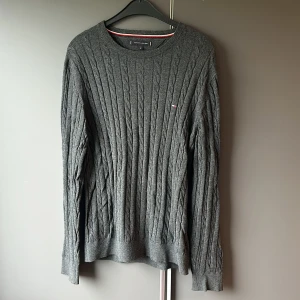 Grå kabelstickad tröja från Tommy Hilfiger - Säljer en snygg grå kabelstickad tröja från Tommy Hilfiger. Den är gjord av premium bomull och ull, vilket gör den både varm och bekväm. Tröjan har långa ärmar och en klassisk rund halsringning. Perfekt för höst och vinter! Den är i mycket bra skick!