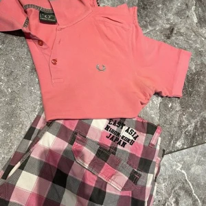 Polotröja piké fred Perry  - En rosa Fred Perry polotröja med grå detaljer runt kragen och knapparna. Shortsen är i rosa, svart och vitt rutmönster med fickor på sidorna. Köpt utomlands