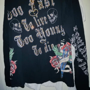 Svart långärmad tröja med dödskalletryck - Säljer en svart långärmad tröja med coolt tryck av dödskallar och texten 'Too Fast to Live, Too Young to Die'. Tröjan har även detaljer med rosor och en dolk. Perfekt för en rockig stil eller konsertkvällar. Den är i bra skick och redo för nya äventyr!