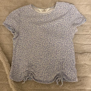 Blommig topp från H&M - Säljer en söt blommig topp från H&M i ljusblått med vita blommor. Toppen har korta ärmar och en rund halsringning. Den har också fina knytdetaljer på sidorna som ger en extra touch. Perfekt för vår och sommar!🌸🌼