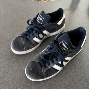 Mörkblå Adidas Campus sneakers - Säljer ett par mörkblå Adidas Campus sneakers i mocka. De har vita detaljer och klassiska tre ränder på sidan. Skorna är välanvända men fortfarande i bra skick. Perfekta för vardagsbruk med en stilren look. Storlek 42. Nypris: 1145 kr.