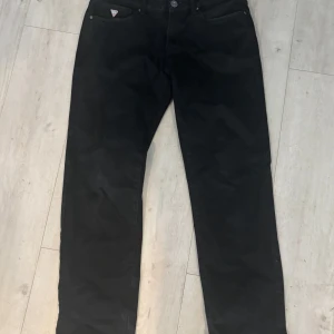 Svarta jeans från Guess - Säljer ett par snygga svarta jeans från Guess. De har en slim straight passform som sitter perfekt för en stilren look. Jeansen är i bra skick och har en klassisk femficksdesign med en diskret logga på bakfickan. Perfekta för både vardag och fest!