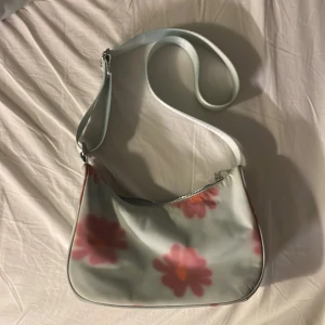 Blommig axelväska från Weekday - Säljer en superfin axelväska från Weekday med ett blommigt mönster i rosa och orange. Väskan har en dragkedja upptill och en justerbar axelrem. Perfekt för att liva upp vilken outfit som helst!
