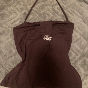 Brun halterneck topp med smyckesdetalj - Säljer en snygg brun halterneck topp med en fin smyckesdetalj framtill. Toppen är perfekt för en utekväll eller fest och har en åtsittande passform som framhäver figuren. Den är axelbandslös med knytning runt nacken, vilket ger en elegant look.