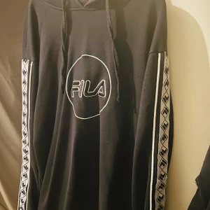 Svart hoodie från FILA - Säljer en svart hoodie från FILA i storlek S. Den har en stor logga på framsidan och coola mönstrade band längs ärmarna. Perfekt för en avslappnad stil och passar bra till både höst och vinter. 