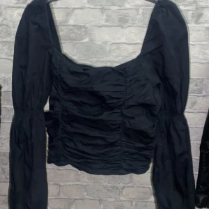 Svart långärmad topp - Säljer en snygg svart topp med långa ärmar och rynkade detaljer. Toppen har en croppad passform och en fyrkantig halsringning som ger en trendig look. Den är i storlek M men funkar som S/XS. Tröjan är helt ny. Perfekt för både vardag och fest!