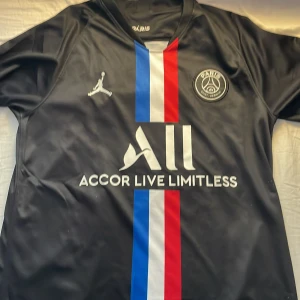 Svart PSG fotbollströja med Mbappé 7 - Snygg svart PSG-tröja med Mbappé 7 på ryggen. Framsidan har Paris Saint-Germain-logga och Jordan-märke. Vertikala ränder i blått och rött ger en cool kontrast. Perfekt för fans av både PSG och Mbappé!