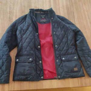 Svart quiltad jacka  - Snygg svart quiltad jacka med röd insida. Jackan har en dragkedja och knappar framtill samt två fickor med knappar. Perfekt för höst och vår!
