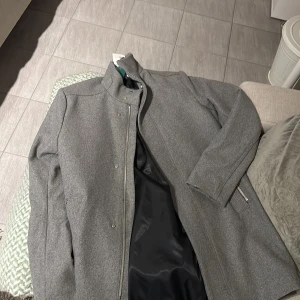 Grå kappa från H&M Conscious - Säljer en stilren grå kappa från H&M Conscious. Kappan är tillverkad av återvunnen ull och har en slät design med knappar framtill. Perfekt för höst- och vårsäsongen.