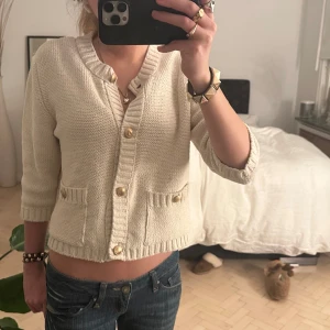 stickad kofta med knappar - Säljer en snygg beige stickad kofta med trekvartsärm och dekorativa knappar framtill. Perfekt för en stilren och bekväm look. Passar bra till jeans eller kjol.