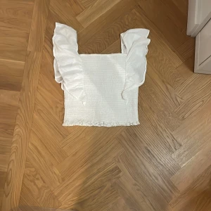 Vit topp från Kappahl - Säljer en söt vit topp från Kappahl med rynkade detaljer och volangärmar. Perfekt för sommarens varma dagar. Toppen har en smickrande passform och är tillverkad i ett bekvämt material.