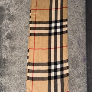 Beige rutig halsduk - Snygg beige halsduk med klassiskt rutigt mönster i svart, vitt och rött. Perfekt för att hålla dig varm och stilren under kyliga dagar. Den har fransar i ändarna som ger en extra touch av elegans.