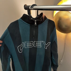 Randig tröja från Obey - Säljer en cool randig tröja från Obey i svart och blått. Tröjan har en dragkedja vid halsen och är långärmad. Perfekt för en avslappnad stil.