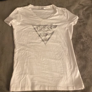 Vit t-shirt från Guess - Snygg vit t-shirt från Guess med det ikoniska triangellogotypen tryckt på framsidan. T-shirten har en v-ringad hals och korta ärmar. Den är i bra skick och har nästan aldrig används.