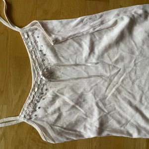 Vit ribbad linnetopp med spetsdetaljer - Säljer en söt vit ribbad linnetopp med spetsdetaljer och små knappar framtill. Toppen har smala axelband och en dekorativ snörning vid halsringningen. Perfekt för varma sommardagar!