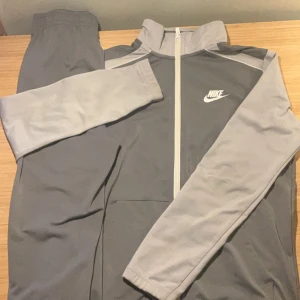 Grå träningsjacka och byxor från Nike - Säljer en grå träningsjacka från Nike med dragkedja och långärmade ärmar. Jackan har en stilren design med Nike-logga på bröstet och är tillverkad i polyester. Perfekt för sport och träning den är varm för vintern men det ändå är att byxorna Nike märke är borta men annars funkar dom perfekt 147-158 på byxorna samt jackan 