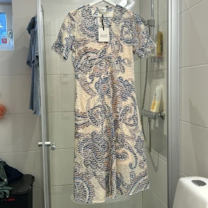 Mönstrad klänning från Second Female - Snygg klänning från Second Female med ett detaljerat mönster i blått och beige. Klänningen har korta ärmar och en v-ringad hals. Perfekt för en stilfull look.