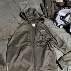 Svart Therma-Fit hoodie från Nike - Säljer en svart Therma-Fit hoodie från Nike med dragkedja och huva. Perfekt för kyliga dagar med sin värmeisolerande design. Den har långa ärmar och en stilren look med Nike-logga på bröstet.