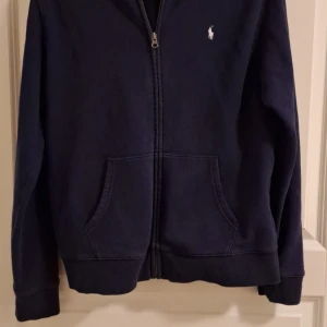 Mörkblå hoodie från Ralph Lauren - Säljer en mörkblå  hoodie från Ralph Lauren med dragkedja och huva. Den har en liten logga på bröstet och text på huvan. Perfekt för en avslappnad stil. Frågor? Skriv direkt!