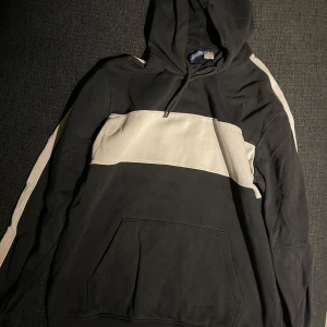 Svart och vit hoodie Divided H&M - Snygg svart hoodie från H&M, med vit rand över bröstet och längs ärmarna. Tröjan har en praktisk ficka framtill och en bekväm huva. 