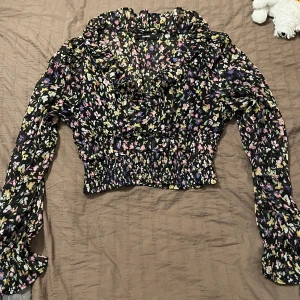 Blommig blus med volangdetaljer - Säljer en söt blommig blus med volangdetaljer och långa ärmar. Blusen har en elastisk midja och är perfekt för både vardag och fest. Den har ett färgglatt mönster med inslag av rosa, gul och lila på en svart bakgrund.💕