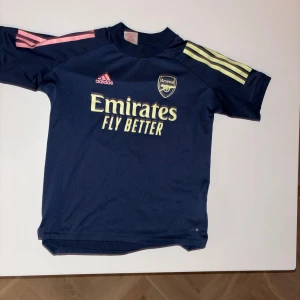 Mörkblå Arsenal fotbollströja från Adidas - Snygg mörkblå Arsenal fotbollströja från Adidas med korta ärmar. Tröjan har gula och rosa detaljer samt klubbens emblem.skriv vid frågor!