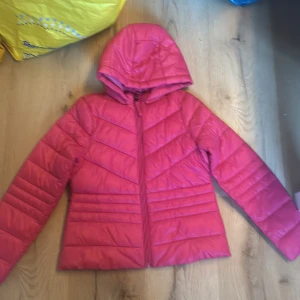 Rosa pufferjacka från Vero Moda - Snygg rosa pufferjacka från Vero Moda med huva och dragkedja. Perfekt för kyliga dagar med sin quiltade design som ger extra värme. Jackan har långa ärmar och en figurnära passform.