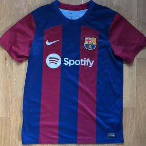 Barcelona fotbollströja  - Snygg fotbollströja från Nike i blått och rött med FCB-logga och Spotify-sponsortryck. Tröjan har korta ärmar och ett klassiskt randigt mönster. Perfekt för fotbollsfans! Inget tryck på ryggen därav pris