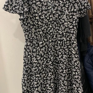 Svart blommig klänning från new yorker  - Charmig svart klänning med vita blommor från new yorker. Den har korta ärmar och en bekväm passform som passar perfekt för både vardag och speciella tillfällen. 
