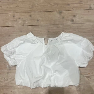 Vit blus från Zara - Säljer en söt vit blus från Zara med puffiga kortärmar och rund hals. Perfekt för en stilren look. Blusen har en knapp vid halsen (bakom) och är tillverkad i ett lätt material.