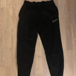Svarta mjukisbyxor från Nike - Säljer ett par svarta mjukisbyxor från Nike med Dri-FIT-teknologi. Byxorna har en elastisk midja med snörning och en liten Nike-logga på benet samt ficka med dragkedja.