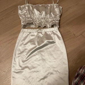 Vit satinlinnetopp med kjol - Snygg vit satinlinnetopp med smala axelband och matchande kjol. Toppen har en elastisk midja med volangdetaljer. Toppen är i S & kjolen i XS. Verkligen Såå fint! Mini fläck på framsidan av kjolen som knappt är märkbar  (se sista bild) 